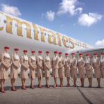 Emirates cabin crew in beige uniforms and red hats posing in front of Emirates-branded Airbus aircraft on tarmac